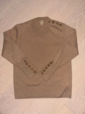 Beige stickad tröja från Burberry - Snygg beige stickad tröja från Burberry med guldfärgade dekorativa knappar på ena axeln och vid ärmsluten. Tröjan har rund halsringning och ribbade muddar. Tillverkad i mjukt material som känns lyxigt och stilrent. Storlek L men passar (S)
