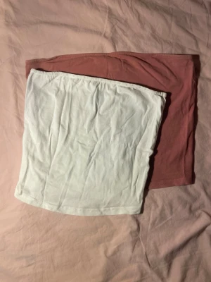 Två tubtoppar i vitt och rosa (XS/S) - Säljer två bandeau tubtoppar, en i vit och en i ljusrosa. Båda är ärmlösa och har en enkel, rak passform. Perfekta att styla till jeans eller kjol för en clean och enkel look. Materialet känns mjukt och stretchigt, troligen bomull eller bomullsblandning. Har tecken på användning. Rosa är S och den vita är XS.