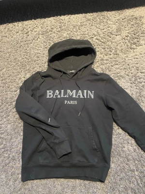 Svart Balmain hoodie med logga - Cool svart hoodie från Balmain med stor vit logga 'BALMAIN PARIS' på bröstet. Klassisk huva med dragsnören och en stor magficka. Mjukt material och ribbade muddar vid ärmslut och nederkant. Perfekt för dig som gillar exklusiva streetwear-vibes.