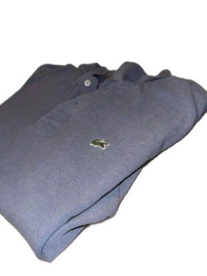 Blå pikétröja från Lacoste - Storlek L Passar M / L Klassisk blå pikétröja från Lacoste med den ikoniska gröna krokodilloggan på bröstet. Tröjan har krage, knappar framtill och är tillverkad i mjuk bomull. Perfekt för en clean och stilren look.