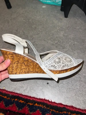 Vita kilklackssandaler med spets - Säljer ett par vita sandaler från Tamaris med kilklack i trälook. Ovandelen har fina spetsdetaljer och glittrande stenar på remmarna. Sandalerna har öppen tå och justerbar rem runt hälen. Perfekta för dig som vill ha både höjd och stil.