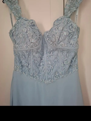 Ljusblå balklänning med spetsdetaljer - Jättefin ljusblå långklänning med spets axelband som hänger ner lite på armen och vackra spetsdetaljer över bysten. Klänningen har blommigt broderi upptill och en flytande kjol i ett lätt material. Jag köpte den på Carolina bröllop, bal och gala i Vara för 5300kr och använd endast på studentbalen och har varit på kemtvätt efter det, inga anmärkningar finns