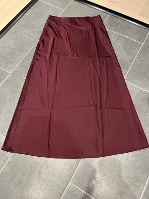 Vinröd långkjol i satin - Snygg vinröd långkjol i satin med lätt utsvängd passform. Kjolen har en slät midja och ett stilrent, minimalistiskt uttryck. Perfekt för dig som gillar en elegant och enkel look. Liten fläck bild 3