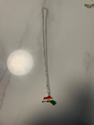 Halsband med Kurdistan-hänge - Snyggt halsband med ett hänge i form av Kurdistan, dekorerat med röd, vit och grön emalj samt en gul sol i mitten. Kedjan är silverfärgad och har en enkel, stilren design. Perfekt accessoar för att visa din identitet och stil.