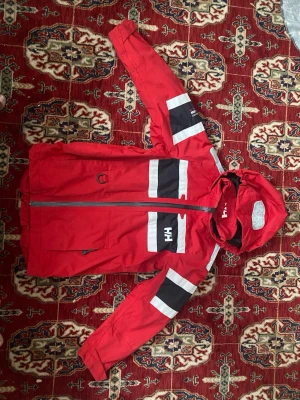 Röd Helly Hansen vindjacka med huva - Snygg röd vindjacka från Helly Hansen med huva och dragkedja. Jackan har svarta och vita detaljer på ärmar och bröst, samt HH-logga på bröst och ärm. Perfekt för att skydda mot vind och regn, med praktiska fickor och sportig design.
