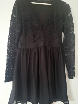 Svart spetsklänning från Nelly L/40 - Snygg svart kortklänning med spetsdetaljer och v-ringning. Klänningen har långärmade spetsärmar och ett flowy kjolparti i tunt tyg. Perfekt för dig som gillar en feminin och elegant stil. Ingen synlig logga. Innerklänning.
