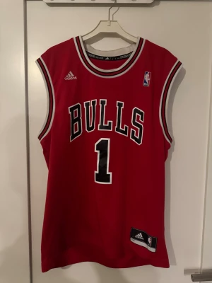 Chicago Bulls basketlinne Adidas - Rött Chicago Bulls basketlinne från Adidas med nummer 1 och namnet Rose på ryggen. Klassisk NBA-design med vita och svarta detaljer runt hals och ärmhål. Tillverkat i lätt och ventilerande material, perfekt för basket eller streetwear.