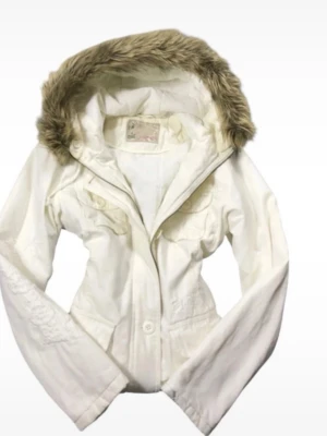 Vit parkasjacka med fuskpäls - Snygg vit parkasjacka med huva och fuskpäls runt kanten. Jackan har två bröstfickor med lock och knappar samt dragkedja framtill. Perfekt för kyliga dagar och ger en fräsch look.