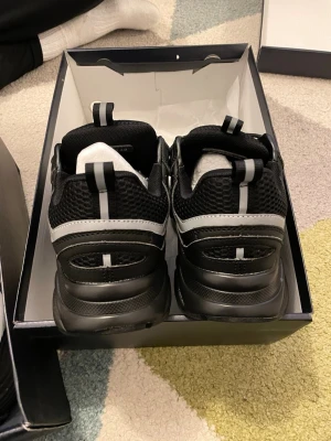 Svarta D**r b22 med grå detaljer - Snygga svarta sneakers med grå och vita detaljer. Ovandelen är i mesh och syntetmaterial för bra andningsförmåga och komfort. Skorna har en sportig look med reflekterande detaljer och klassisk snörning. Perfekta för dig som gillar stilrena och moderna sneakers.