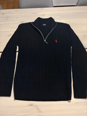 Mörkblå kabelstickad tröja Polo Ralph Lauren - Säljer en mörkblå kabelstickad tröja från Polo Ralph Lauren med halv dragkedja och röd broderad logga på bröstet. Tröjan har hög krage och långa ärmar, perfekt för lager på lager. Klassisk och stilren design i mjukt material, är ej säker på om den är äkta, står L på taggen men passar S-M bättre, pris går att diskutera vid snabb affär!