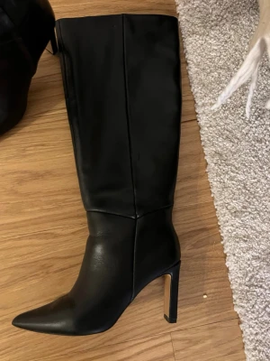 Svarta höga boots i skinn från NA-KD - Stilrena svarta boots i äkta skinn från NA-KD med högt skaft och spetsig tå. Skorna har en stadig klack och dragkedja på insidan för enkel påtagning. Perfekta för att lyfta din outfit med en edgy vibe. Klacken är 8 cm
