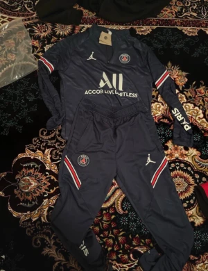 PSG träningsset i mörkblått - Snyggt träningsset från PSG och Jordan i mörkblått med vita och röda detaljer. Setet består av en långärmad tröja med dragkedja och matchande byxor, båda med PSG-logga och Jordan-logga. Materialet är troligtvis polyester, perfekt för träning eller sport.