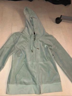 Ljusgrön velourhoodie med dragkedja - Mjuk ljusgrön hoodie i velour från Juicy Couture med dragkedja framtill, fickor och broderad logga på bröstet. Klassisk huva med dragsko och ribbade muddar. Med matchande byxor till. Perfekt för en chill och avslappnad stil. Tröjan har några fläckar på sig. Vet inte om dom går att få bort.