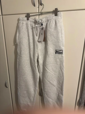 Grå mjukisbyxor från Stussy - Säljer ett par ljusgrå mjukisbyxor från Stussy med logotyp på vänster ben. Byxorna har elastisk midja med dragsko, sidofickor och ribbade benslut. Perfekta för chill och streetwear. Materialet är mjukt och bekvämt, troligtvis bomullsblandning. OBS TAGG SÄGER XXL MEN DET STÄMMER INTE. PASSAR M-L