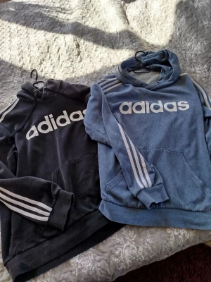 Adidas set: hoodies & tights - Två Adidas huvtröjor, en svart och en blå, båda med vita ränder på ärmarna och stor logga framtill. Tillhörande svarta tights, ett par långa och ett par 3/4-långa, båda med klassiska vita ränder längs benen. Perfekt för träning eller chill. Materialet är mjukt bomullsmix.