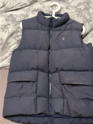 Mörkblå dunväst från GANT - Snygg mörkblå dunväst från GANT med hög krage och dragkedja framtill. Västen har två stora fickor med lock och diskret GANT-logga på bröstet. Perfekt för lager på lager och en clean look.