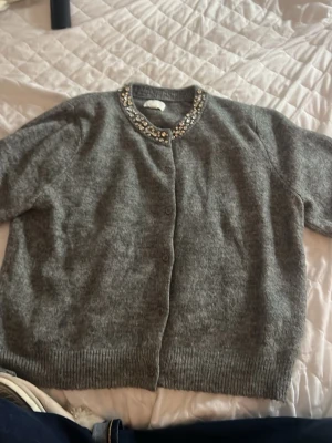 Grå kofta med strassdetaljer - Mysig grå kofta från H&M med rund halsringning och glittrande strassdetaljer runt kragen. Koftan har knäppning framtill och ribbade muddar vid ärmslut och nederkant. Perfekt för dig som vill ha något extra till din outfit.