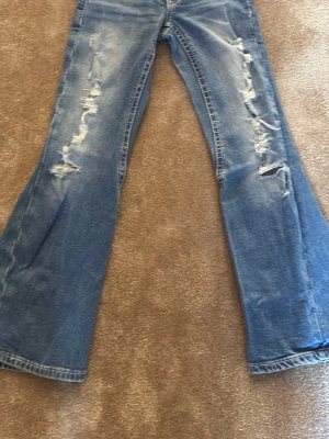 Blå slitna bootcut jeans - Säljer ett par blå bootcut jeans med slitningar och hål på framsidan. Jeansen har utsvängda ben och klassiska bakfickor med knappdetaljer. Perfekta för en avslappnad och trendig look. Tillverkade i denim.