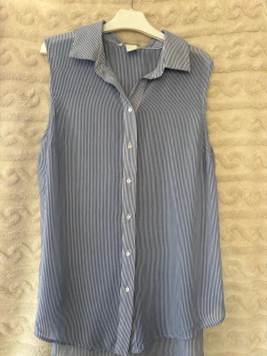 Blåvit ärmlös randig blus - Snygg ärmlös blus med tunna blå och vita ränder, klassisk krage och knäppning framtill. Blusen har en avslappnad passform och är tillverkad i ett lätt, luftigt tyg som passar perfekt till sommaren. Enkel att styla med jeans eller kjol.