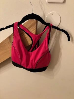 Rosa sport-bh med svarta detaljer - Snygg sport-bh i stark rosa färg med svarta kontrastsömmar och bred svart resår nedtill. Bh:n har brottarrygg och justerbar knäppning bak för extra stöd!❣️storlek S men sitter mer som en xs