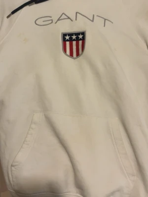 Vit hoodie från GANT med logga - Säljer en vit hoodie från GANT med broderad logga och amerikanskinspirerat sköldmärke på bröstet. Hoodien har huva med mörka snören och en stor magficka framtill. Tillverkad i mjukt bomullsmaterial, perfekt för en avslappnad och stilren look.
