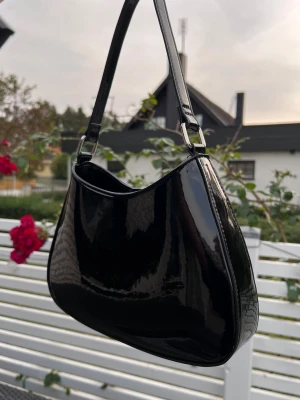 Glansig y2k väska i svart lack, Gina Tricot - Snygg svart vädka i glansigt lackmaterial med silverfärgade detaljer. Väskan har en mjukt rundad form och stängs med magnetknapp. Använd två gånger. Nypris 349kr. Längd: 28,5 cm - Höjd: 17 cm - Band: 29 cm