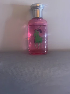 Rosa parfym från Ralph Lauren - En snygg parfymflaska i rosa glas från Ralph Lauren med det klassiska gröna polospelar-motivet på framsidan. Flaskan har en rundad form och ett silverfärgat lock som ger en lyxig känsla. Perfekt för dig som gillar stilrena och färgglada detaljer.