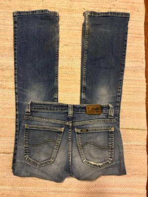 Blå bootcut jeans från Lee - Midjemått =36 cm innerbenslängd = 76 cm. Snygga 90-tals blå bootcut jeans från Lee med slitningar på framsidan och klassiska bakfickor med Lee-logga. Jeansen har låg midja och är tillverkade i denim med lätt tvättad look. Har ett hål i skrevet som dock går att sy ihop jätte enkelt för hand el med symaskin