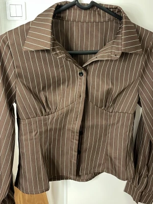 Brun randig skjorta med knappar - Snygg brun skjorta med vita vertikala ränder och markerad krage. Skjortan har långa ärmar, figurnära passform och knäppning framtill. Perfekt för dig som gillar en trendig och stilren look.