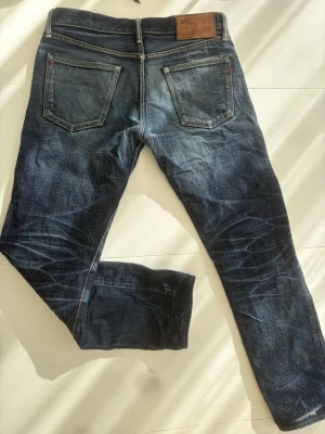 Selvedge jeans - Säljer ett par mörkblå selvedge jeans från naked & famous med slitningar och snyggt tvättad look. Jeansen har raka ben, och knäppning med knappar framtill. Materialet är klassisk denim i bomull. Perfekta för en avslappnad och trendig stil.