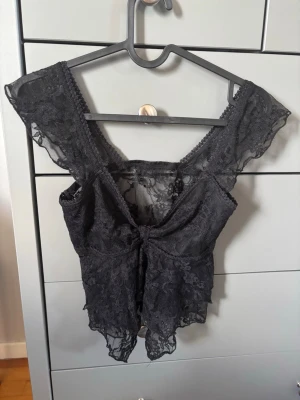 Svart spetsig blus med v-ringning - Snygg svart blus i spets med v-ringning och korta, genomskinliga ärmar. Toppen har en feminin och romantisk look med fina spetsdetaljer och lätt croppad passform. Perfekt att styla med jeans eller kjol för en trendig outfit.