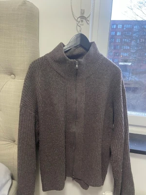 Mörkbrun stickad kofta med dragkedja - Mysig mörkbrun stickad kofta med hög krage och hel dragkedja framtill. Koftan har en ribbad struktur och långa ärmar, perfekt för lager på lager under kyliga dagar.