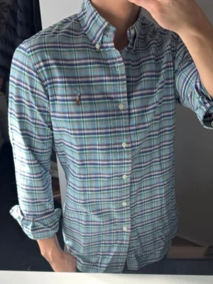 Rutig skjorta från Polo Ralph Lauren - Klassisk rutig skjorta från Polo Ralph Lauren i blå, grön och vit färg. Skjortan har lång ärm, knappar framtill och button-down krage. Tillverkad i mjuk bomull och har den ikoniska broderade loggan på bröstet. Perfekt för en stilren och avslappnad look.