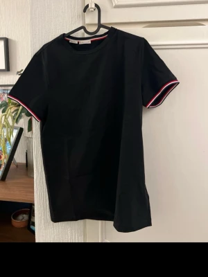 Svart Moncler t-shirt med rödvit kant - Snygg svart t-shirt från Moncler med korta ärmar och coola rödvita detaljer vid ärmsluten. Klassisk rund halsringning och Moncler-logga på ena ärmen. Perfekt till jeans eller shorts för en clean och sportig look.
