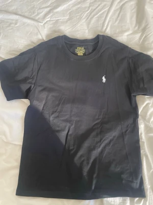 Svart t-shirt från Polo Ralph Lauren - Klassisk svart t-shirt från Polo Ralph Lauren med vit broderad logga på bröstet. Modellen är slim fit. Det skrynkliga går att ta bort med strykjärn aldrig använd.