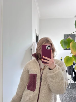 Beige teddyjacka med vinröda detaljer - Mysig beige teddyjacka med hög krage och vinröda detaljer på dragkedja och ficka. Jackan har en oversized passform och är tillverkad i fluffigt syntetmaterial. Perfekt för kalla dagar när du vill vara både varm och snygg.