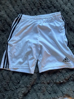 Vita Adidas shorts med svarta ränder - Snygga vita shorts från Adidas med klassiska svarta ränder längs sidorna och Adidas-logga på ena benet. Elastisk midja för skön passform. Perfekta för sport eller chill.