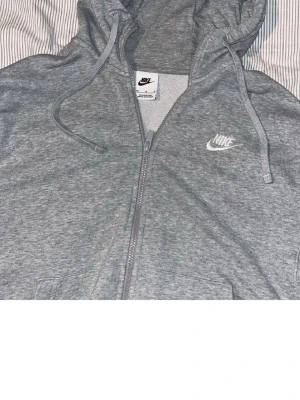 Grå zip hoodie från Nike - Säljer en klassisk grå zip hoodie från Nike med vit broderad logga på bröstet. Hoodien har dragsko i huvan, fickor framtill och är tillverkad i mjuk bomullsblandning. Perfekt för en avslappnad och sportig stil.