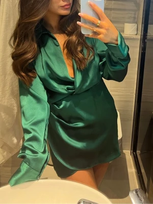 Grön satinklänning  - Snygg grön kort klänning i satin med djup v-ringning och långa ärmar. Klänningen har en avslappnad passform och en elegant, glansig finish som ger en lyxig känsla. Perfekt för dig som vill sticka ut med stil.