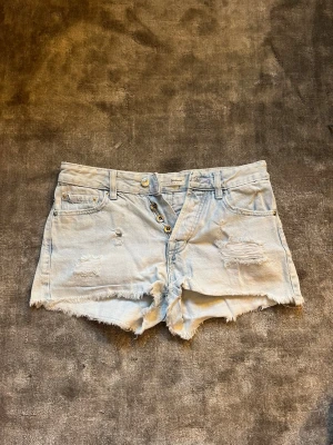 Ljusblå jeansshorts från stradivarius - Säljer ett par ljusblå jeansshorts från stradivarius med fransig kant och slitna detaljer framtill. Shortsen har knappar i gylfen, klassiska fem fickor och ett lädermärke bak i linningen. Perfekta för sommaren! Lågmidjade. Passar 36