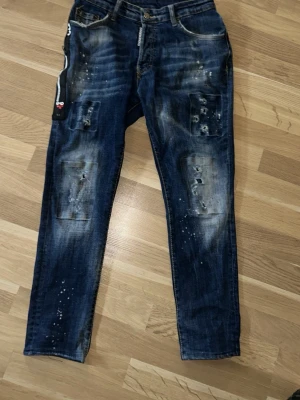 Blå slitna jeans med patchdetaljer - Säljer ett par blå jeans med slitningar, patchade detaljer och färgstänk. Jeansen har en smal passform och klassisk femficksdesign. Materialet är denim i bomull och de har en trendig, vintage look med tydliga slitningar på benen.
