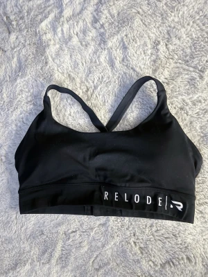 Svart sport-bh från Relode - Snygg svart sport-bh från Relode med korsade axelband och justerbar knäppning bak. Tillverkad i stretchigt funktionsmaterial som ger bra stöd under träningen. Logga framtill i vitt. Perfekt för gym eller löpning 🏋🏼‍♀️🖤