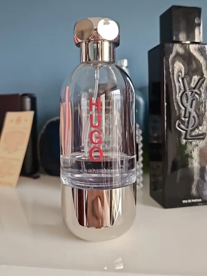 Hugo Boss Element parfym - Modern parfym från Hugo Boss med genomskinlig glasflaska och silverfärgad botten och kork. Flaskan har röd text och en stilren, futuristisk design som sticker ut på hyllan. Perfekt för dig som gillar en fräsch och unik doft.