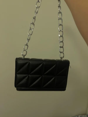 Svart quiltad handväska med kedja - Snygg svart handväska i quiltat mönster med en grov silverfärgad kedja som axelrem. Väskan har en rektangulär form och är tillverkad i konstläder. Perfekt accessoar för att lyfta din outfit.