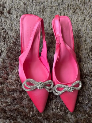 Rosa pumps med strassrosett ASOS - Säljer ett par knallrosa pumps från ASOS med spetsig tå och öppen häl. Skorna har en elegant rosett framtill dekorerad med glittrande strass och är tillverkade i satinliknande material. Perfekta för dig som vill sticka ut med färg och detaljer.