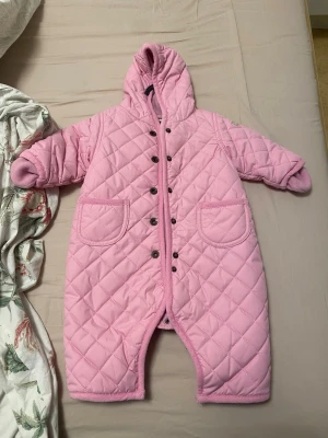 Rosa quiltad overall med huva - Söt rosa quiltad overall med huva och tryckknappar framtill. Overallen har två stora fickor och mjukt fleecefoder på insidan. Perfekt för kyligare dagar och enkel att ta på tack vare knapparna hela vägen ner.