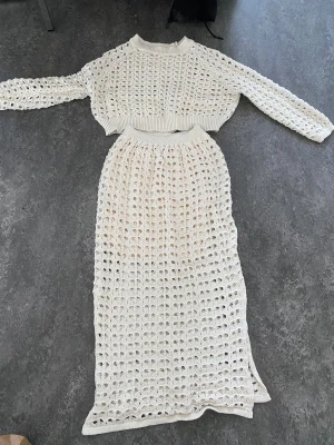 Beige stickat set med kjol och tröja - Säljer ett beige stickat set med långärmad, croppad tröja och matchande lång kjol. Båda delarna har ett öppet virkat mönster och breda ribbade muddar. Kjolen har slits i sidan och resår i midjan. Perfekt för dig som gillar trendiga och luftiga plagg.