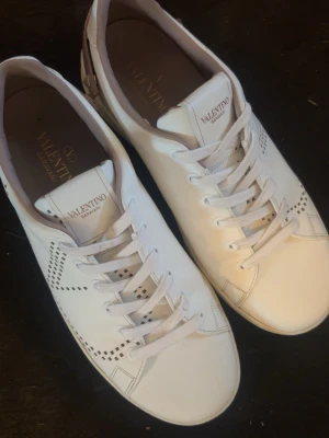 Vita Valentino Garavani sneakers - Snygga vita sneakers från Valentino Garavani i läder med diskreta perforerade detaljer på sidorna och klassisk snörning. Skorna har en låg profil och stilren design som passar perfekt till streetwear och trendiga outfits.