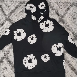 Denim Tears hoodie - Svart hoodie från Denim Tears med stora vita blommönster över hela tröjan. Tröjan har huva och en klassisk magficka framtill. Tillverkad i USA och gjord i mjuk bomull, perfekt för dig som gillar streetwear med unik design.