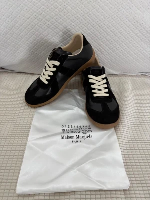  svarta sneakers - Maison Margiela sneakers i svart mocka och läder med vita skosnören och klassisk gummisula. Snygg låg modell med stilren design och beige innerfoder. Perfekta för dig som gillar minimalistisk och trendig stil.